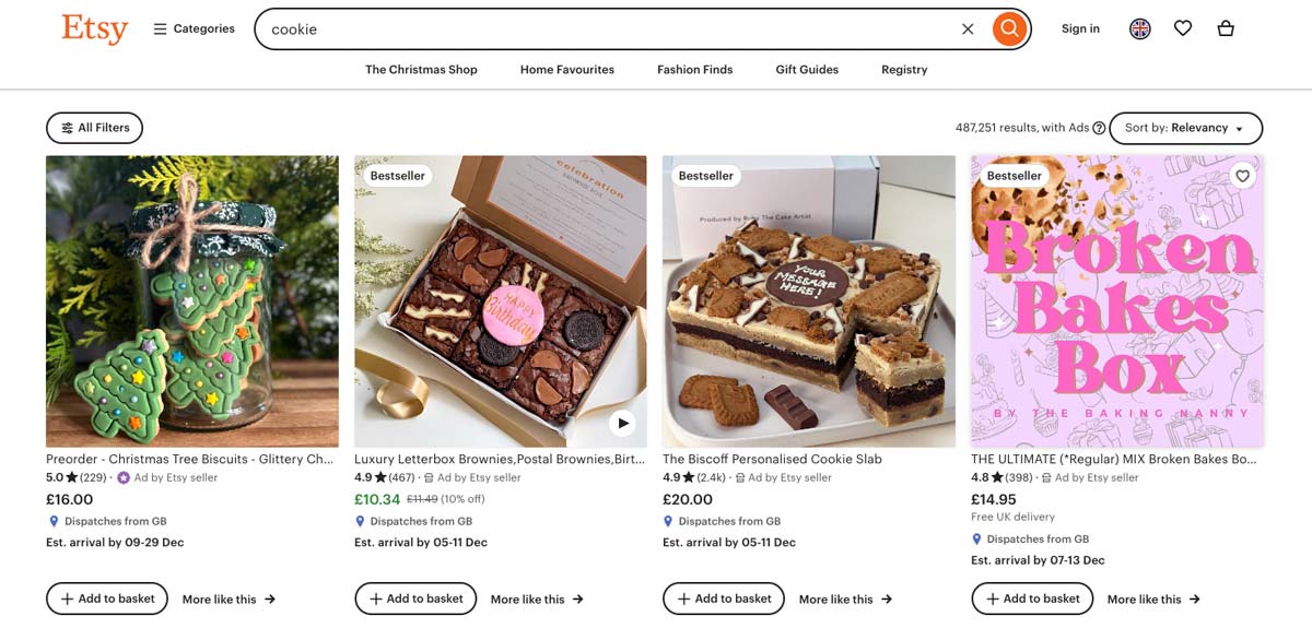 A screenshot of the Etsy website displaying various cookie products for sale, including Christmas tree biscuits in a jar, luxury brownies in a box, a Biscoff personalized cookie slab, and a mixed broken bakes box.